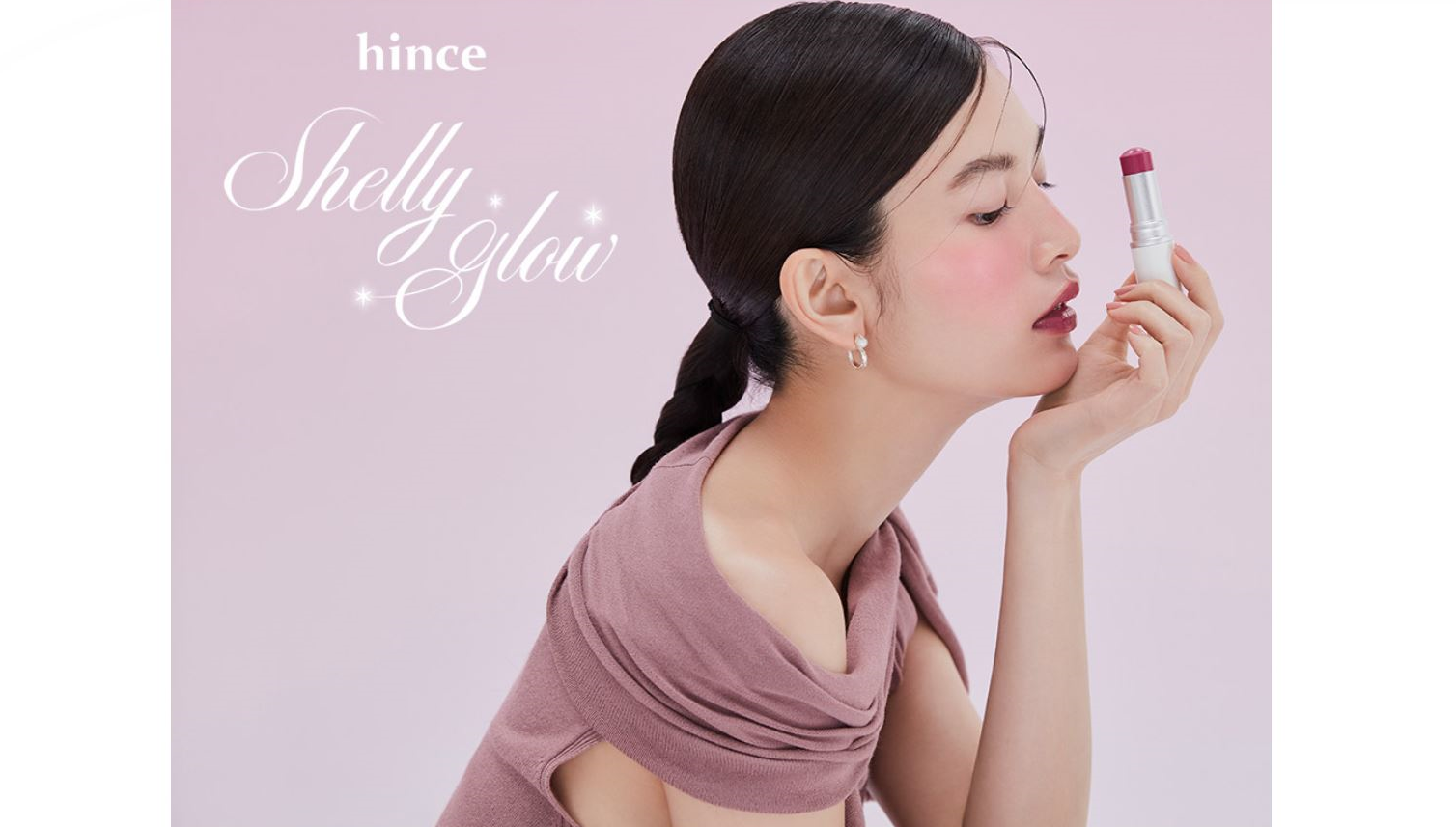Woman applying lipstick with 'hince Shelly Glow' branding on a pink background